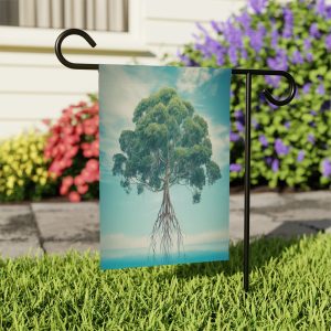 A garden flag with a large green tree on a blue background hangs from a black metal flagpole with a curved tip. The roots of the tree are visible and extend to the bottom of the flag. The flag hangs in a garden with cobblestone pavement and colorful flowers, and in the background a white house with a porch can be seen.